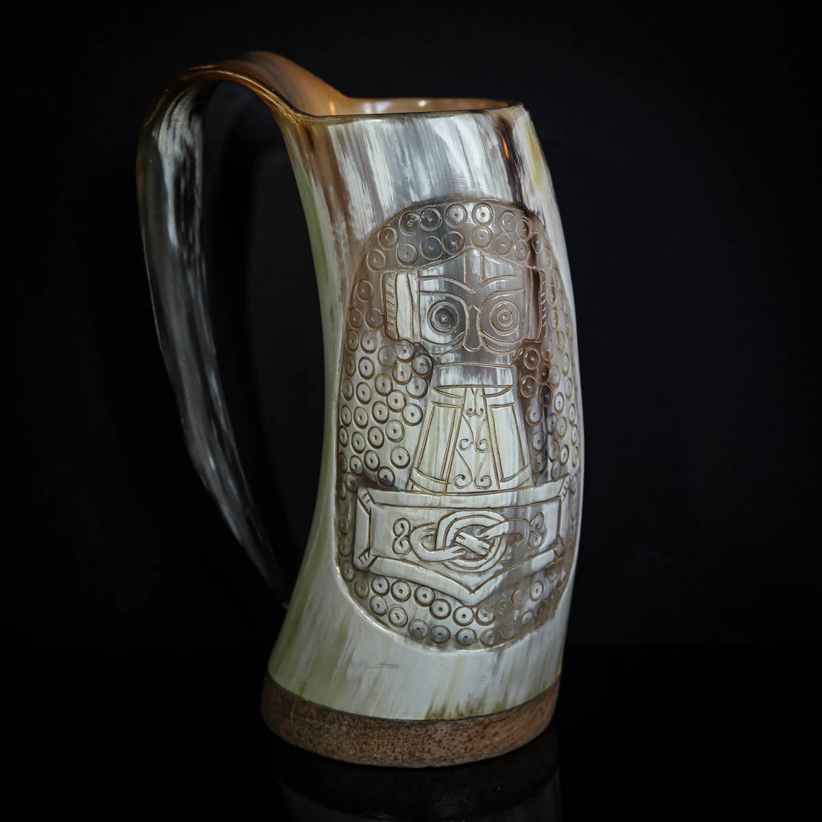 Frontvy av handsnidad hornmugg med detaljerad relief. Tors hammare (Mjölnir) dominerar designen, omgiven av vikingatida knotverk och en krigaransiktsrelief. Mässingsbandet runt basen och det naturliga hornets krämfärgning är synliga.
