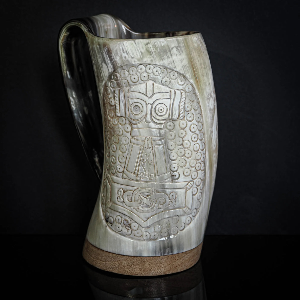 Frontvy av handsnidad hornmugg med komplext snidningsverk. Tors hammare och krigaransiktet dominerar designen med omgivande vikingatida mönster. Mässing, läder och naturligt horn bildar en autentisk Viking-dryckeskärl. Alla detaljer framlysta av professionell studiofotografering.