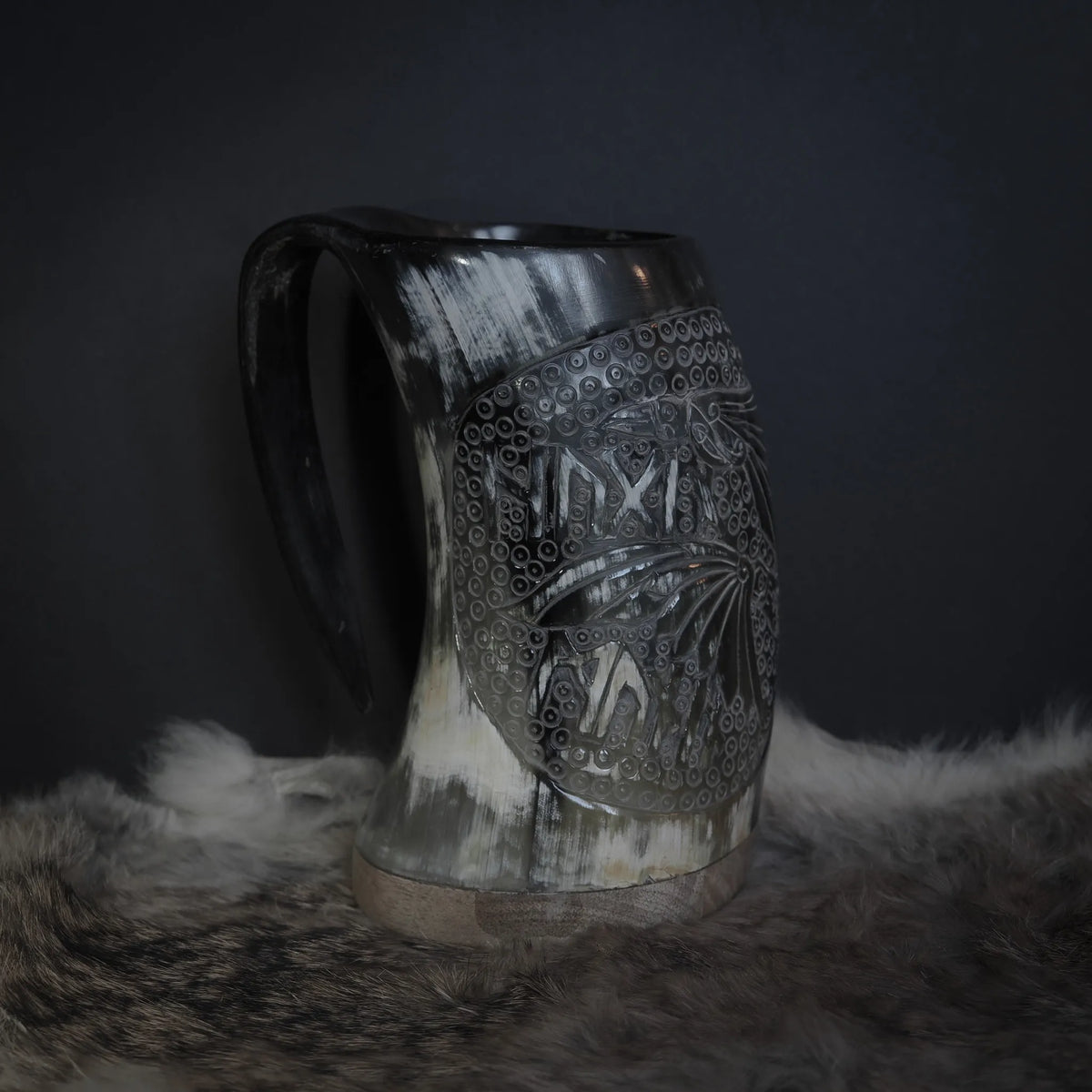 Handsnidad hornmugg med ravnmotiv föreställande Hugin och Munin. Två ravarnas huvuden med intrikat fjädersnidning och uttrycksfulla blickar. Bakgrund fylld med cirkulär prickad mönsterstruktur som representerar ringmössa eller dekorativ infyllnad. Naturligt horn i ljus kräm/vit färg. Kopparband vid basen. Atmosfärisk fotografering med päls i förgrund.