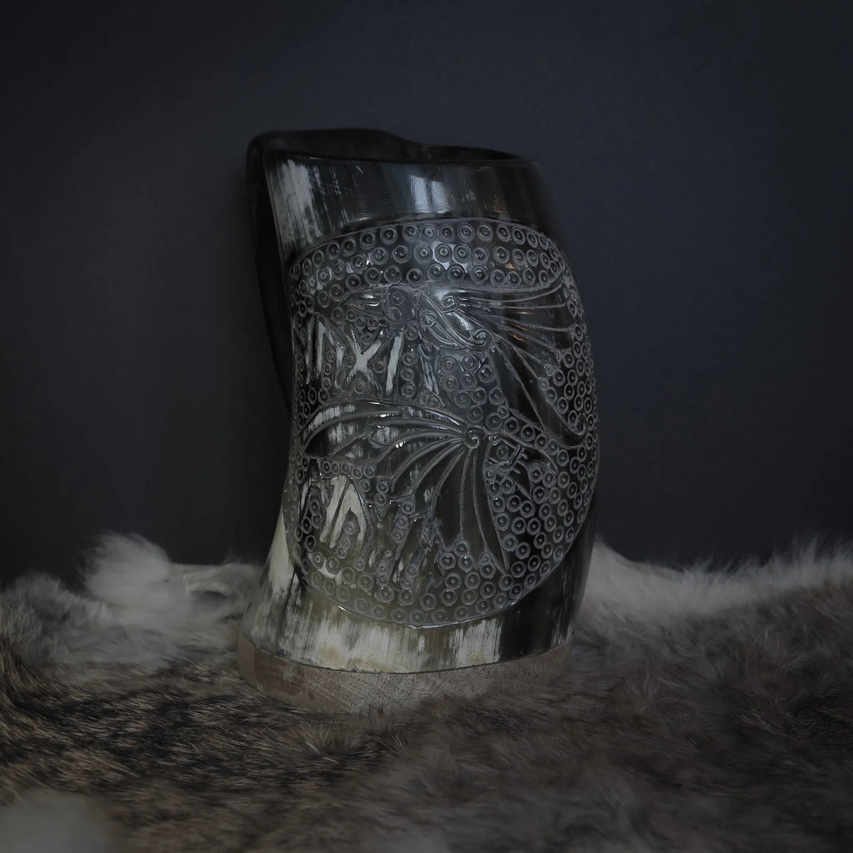 Handsnidad hornmugg med detaljerad ravnmotiv visar två ravarnas huvuden med naturalistisk expression. Fyra vikarnas ögon är animerade och uttrycksfulla. Bakgrunden fylls med cirkulär ringmönsterstruktur som inspireras av ringmössa. Naturligt horn i ljus kräm-vit färg. Kopparband vid basen. Professionell belysning framhäver sniddetaljerna och hornets naturliga kornstruktur.