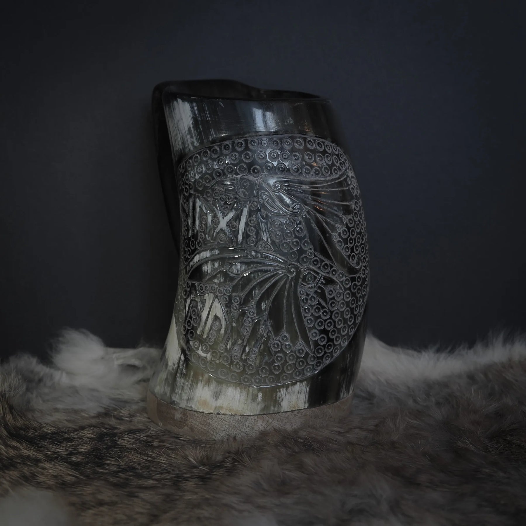 Handsnidad hornmugg med detaljerad ravnmotiv visar två ravarnas huvuden med naturalistisk expression. Fyra vikarnas ögon är animerade och uttrycksfulla. Bakgrunden fylls med cirkulär ringmönsterstruktur som inspireras av ringmössa. Naturligt horn i ljus kräm-vit färg. Kopparband vid basen. Professionell belysning framhäver sniddetaljerna och hornets naturliga kornstruktur.