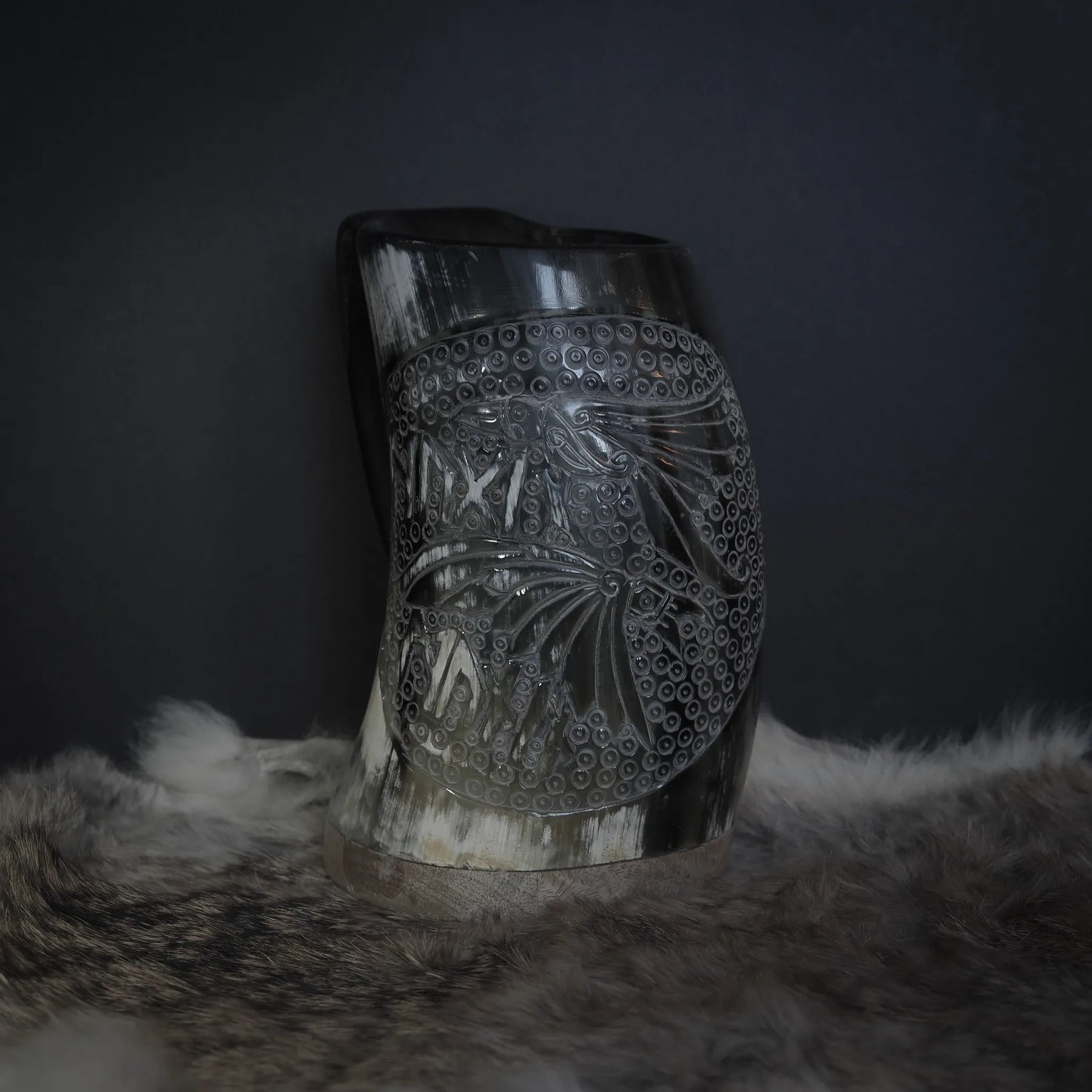 Handsnidad hornmugg av ekologiskt horn med Hugin och Munin-motiv. Cylindrisk form fotograferad från sidan på mörk bakgrund. Karvade fågelmönster med punktradsmönster och naturlig hornets kornstruktur är tydlig.
