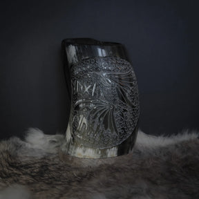Handsnidad hornmugg av ekologiskt horn med Hugin och Munin-motiv. Cylindrisk form fotograferad från sidan på mörk bakgrund. Karvade fågelmönster med punktradsmönster och naturlig hornets kornstruktur är tydlig.