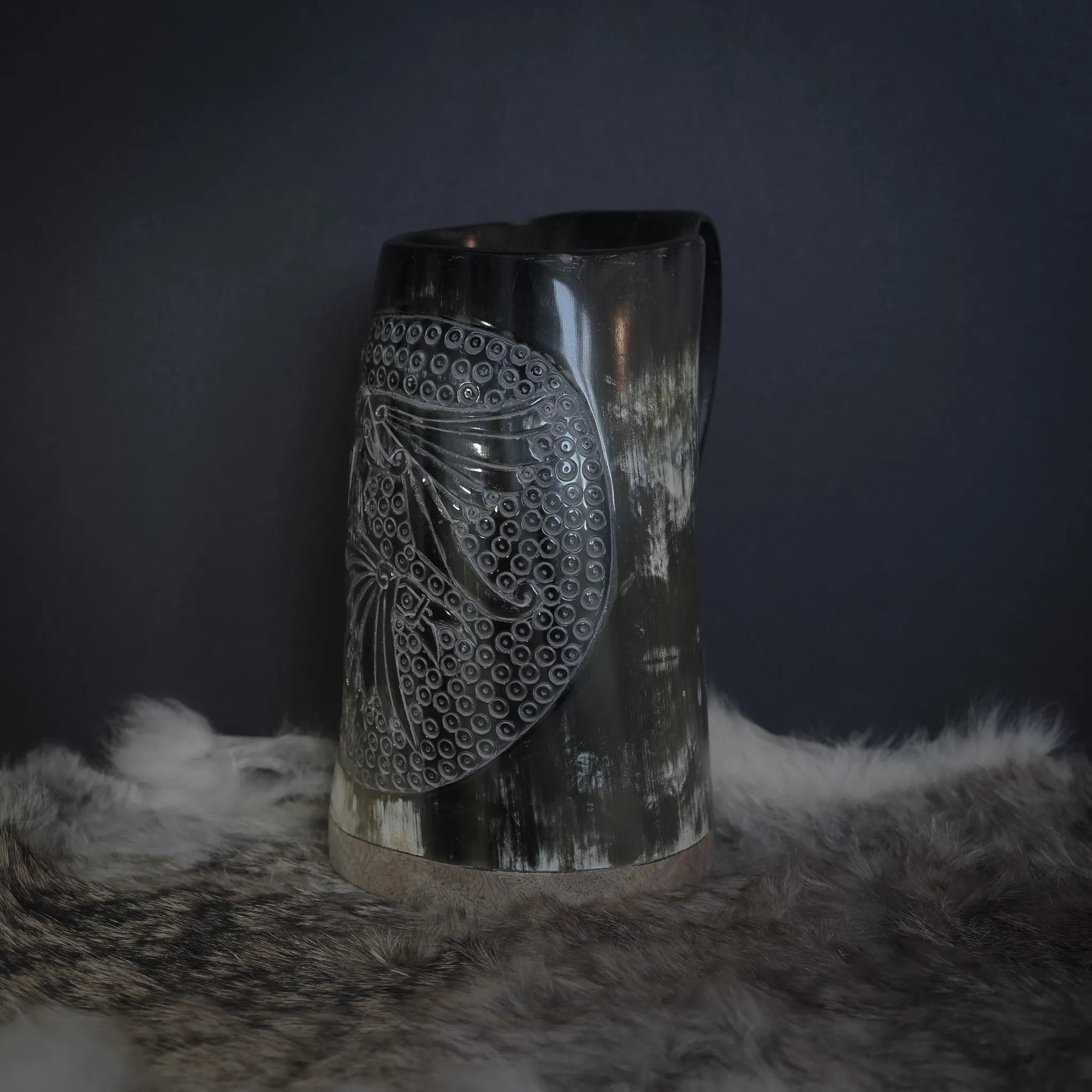 Handsnidad hornmugg fotograferad rakt framifrån. En karvad korpmotif med detaljerade fjädrar omges av ett dekorativt prickmönster. Naturlig hornfärg med markerad kornstruktur.