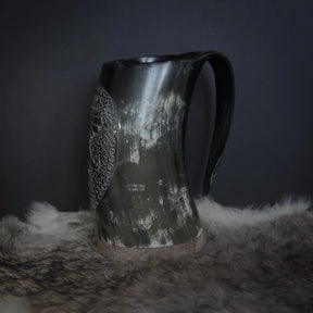 Hornmugg med handkarvad dekor visad i trevärtsvy. Naturlig horn med polerad finish, tydlig handtag och rakkarvad Hugin/Munin-motiv omgiven av prickbroderi.