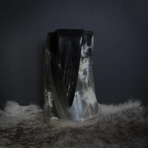 Handkarvad hornmugg visad framifrån. Dekorativ körpfågelmotif med fjäderdetaljer i mitten, omgiven av småprickat mönster. Horn i naturlig färg med ljusa och mörka strimmor.