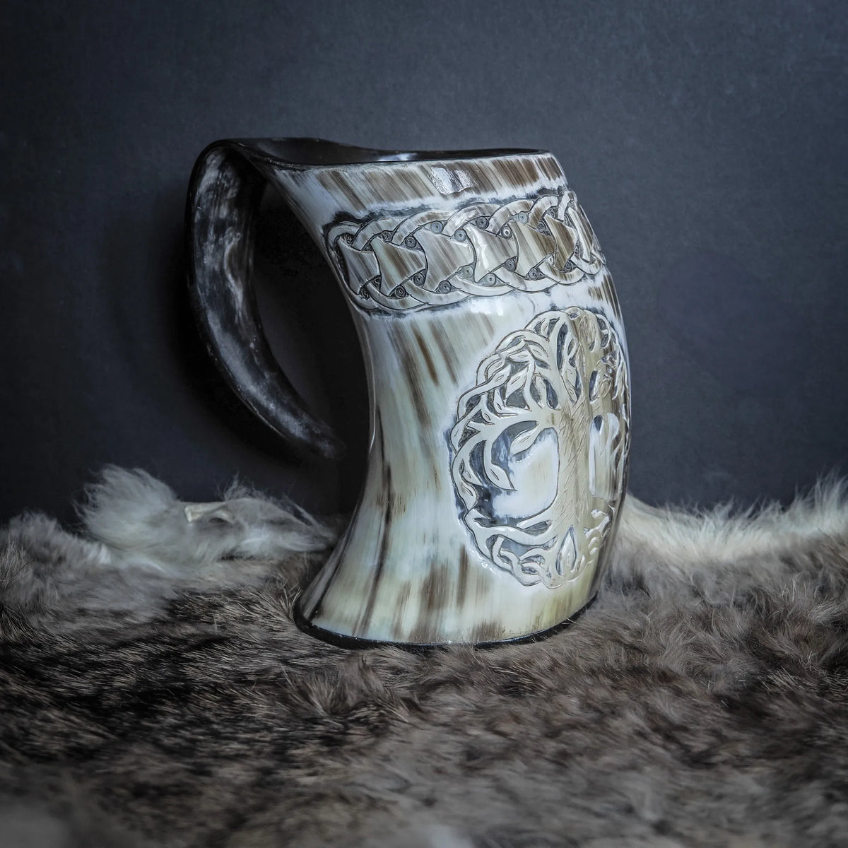 Handsnidad hornmugg i naturligt ljust horn, trevävagvinkel. Gravering av Livets träd med grenar och rötter. Knotband omkring kanten. Handtag synligt. Naturlig hornstruktur och ådror. Djurpälts bakgrund.