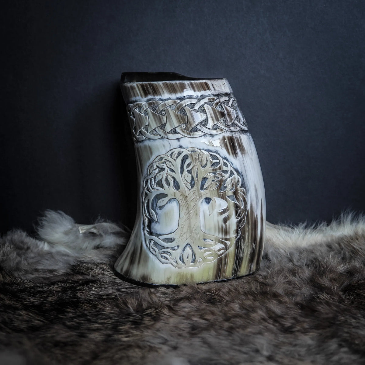 Handsnidad hornmugg i naturellt horn med gravering av Livets träd (Yggdrasil). Visar rötter, grenar och komplicerad trädsignering. Knotband smyckar kanten. Naturlig hornstruktur synlig. Studiofoto på djurpälts bakgrund.