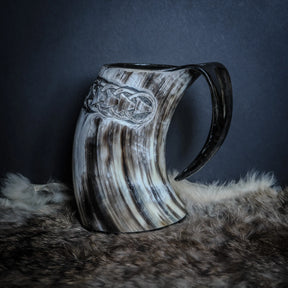 Handsnidad hornmugg med Livets träd-motiv. Naturlig horn i krämfärg och mörkgrå marmor, snidad med vikingtid-knotverk och träd-symbol. Svart hankle. Presenterad på pälsbakgrund.