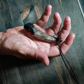 Detaljbild av visselpipan hållen i mänsklig hand, visar munstyckets design och korniga horn-struktur. Vita ben- eller älghorns-tips för kontrast. Svart lädersnöre synligt. Visar den hantverk-gjorda kvaliteten och praktisk storlek.