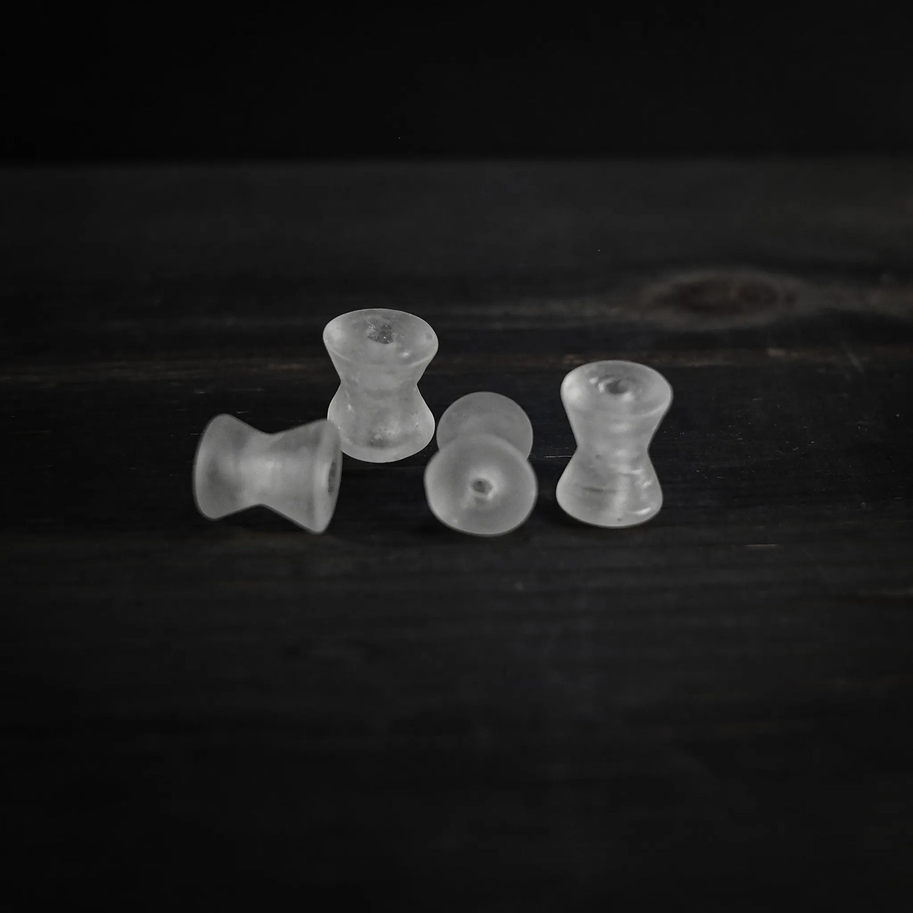 Fyra spolformade pärlor i frostad vitaktig genomskinlig material arrangerade i rad. Varje pärla är cirka 1,7 cm lång och 1,5 cm i diameter med 2 mm genomborrade hål. Studio-produktfotografi mot mörk bakgrund.
