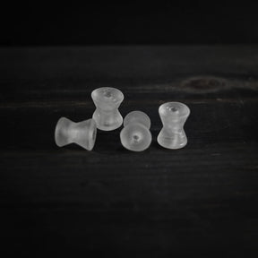 Fyra spolformade pärlor i frostad vitaktig genomskinlig material arrangerade i rad. Varje pärla är cirka 1,7 cm lång och 1,5 cm i diameter med 2 mm genomborrade hål. Studio-produktfotografi mot mörk bakgrund.
