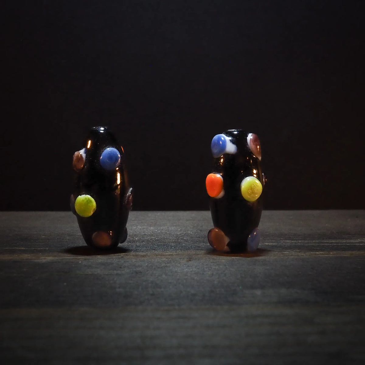 Fyra mörka glaspärlor med ljusa färgade prickar i blå, röd, gul och orange på svart yta omgivna av gröna växtblad