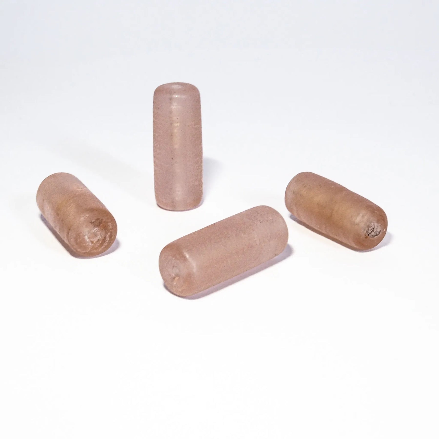 Fyra ljusrosa glaspärlor arrangerade på vit bakgrund. Cylinderformade pärlor med transparent rosaskimrande yta.