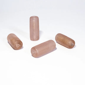Fyra ljusrosa glaspärlor arrangerade på vit bakgrund. Cylinderformade pärlor med transparent rosaskimrande yta.