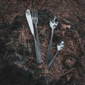 Rostfritt stål bestick med handsmide textur fotograferat på mörkjord och torr gräs i vildmarkssammanhang