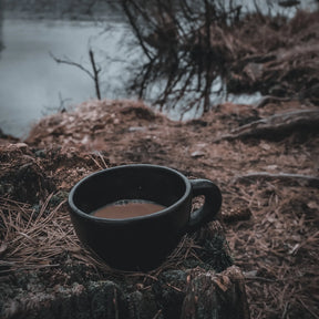 Svart lerkrus med öra fyllt med kaffe, placerad på en väderbitven trädstam vid en sjö. Birka-inspirerad design med glossig svartbränning. Nordisk naturlig miljö med vatten och kärvade grenar i bakgrunden.