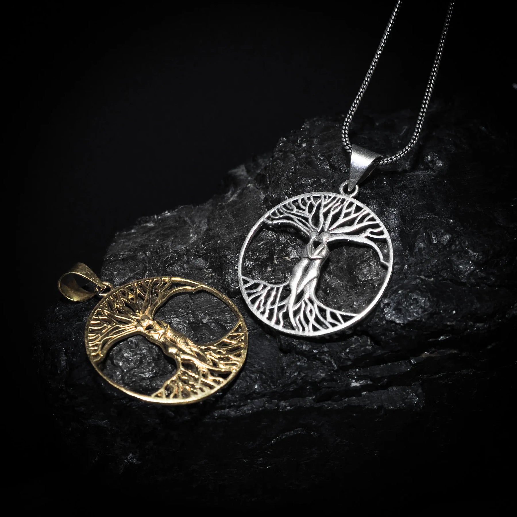 Två träd-amuletter på mörk sten - en i mässing och en i silver - visar samma Yggdrasil-design i olika metallfinisher.