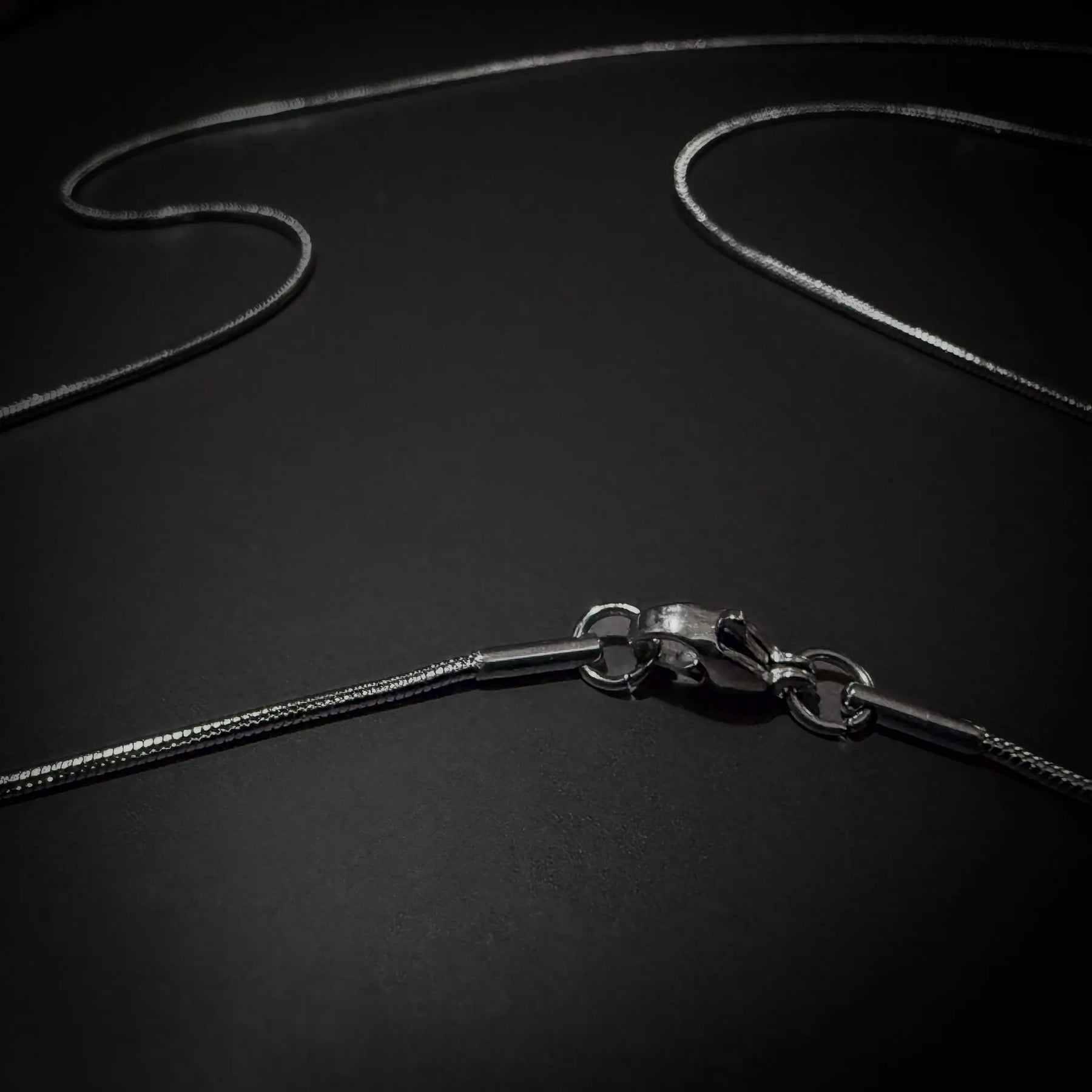 Silverfärgad kirurgisk stålkedja 55cm, 1mm tjock, nickelfri, med slang-mönstrad länkstruktur visad på texturerad yta