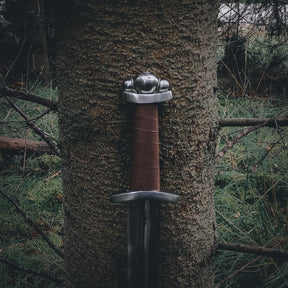 Autentisk vikingsvärd typ S från 800-talet lutat mot mossbeklädd trädstam i skogsomgivning. Kolstålblad med fullerering, läderslaget handtag och bronsbeslag. Stämning av historisk äkthet.
