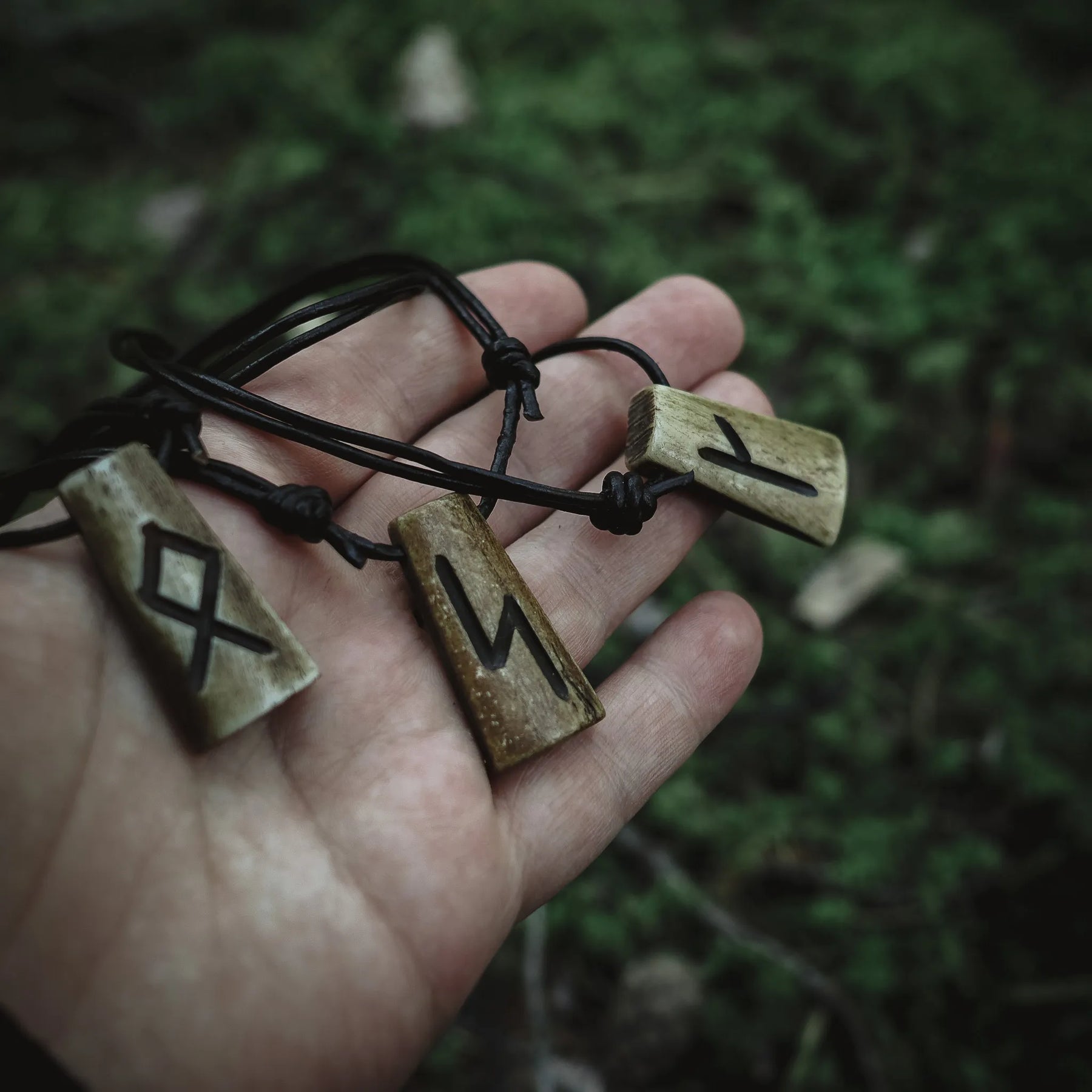 Handgjord runhalsmycke i oxben på svart läderrem. Två rektangulära benmycken med snidade nordiska runor. Fotograferad i hand mot naturlig grön bakgrund. Traditionellt vikingainspirerat smycke.