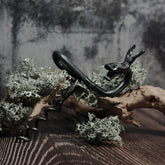 Handsmide älgskruv med älghuvud som handtag i antikpatinerad stålfinish. Positionerad på igenväxt trädgren med mossa, fotograferad mot neutral grå bakgrund med dramatisk belysning. Det spiralformade korkskelutsticket är tydligt synligt.