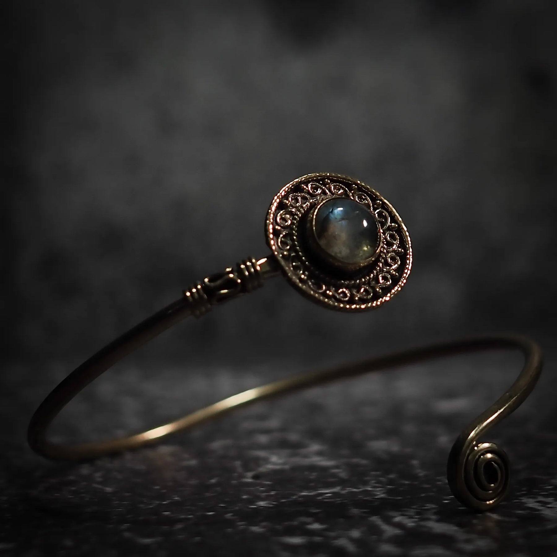 Mässingsarmband med labradorit halvädelssten, ornamentalt filigree-infattning och spiralprydnad på ändar, fotograferat mot mörk bakgrund