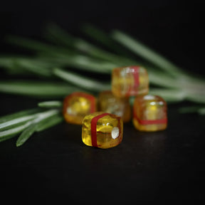 Fem gula kubiska glaspärlor med röda ränder i mjuk fokus mot barrgrenar, visar handgjord glasstruktur och håll