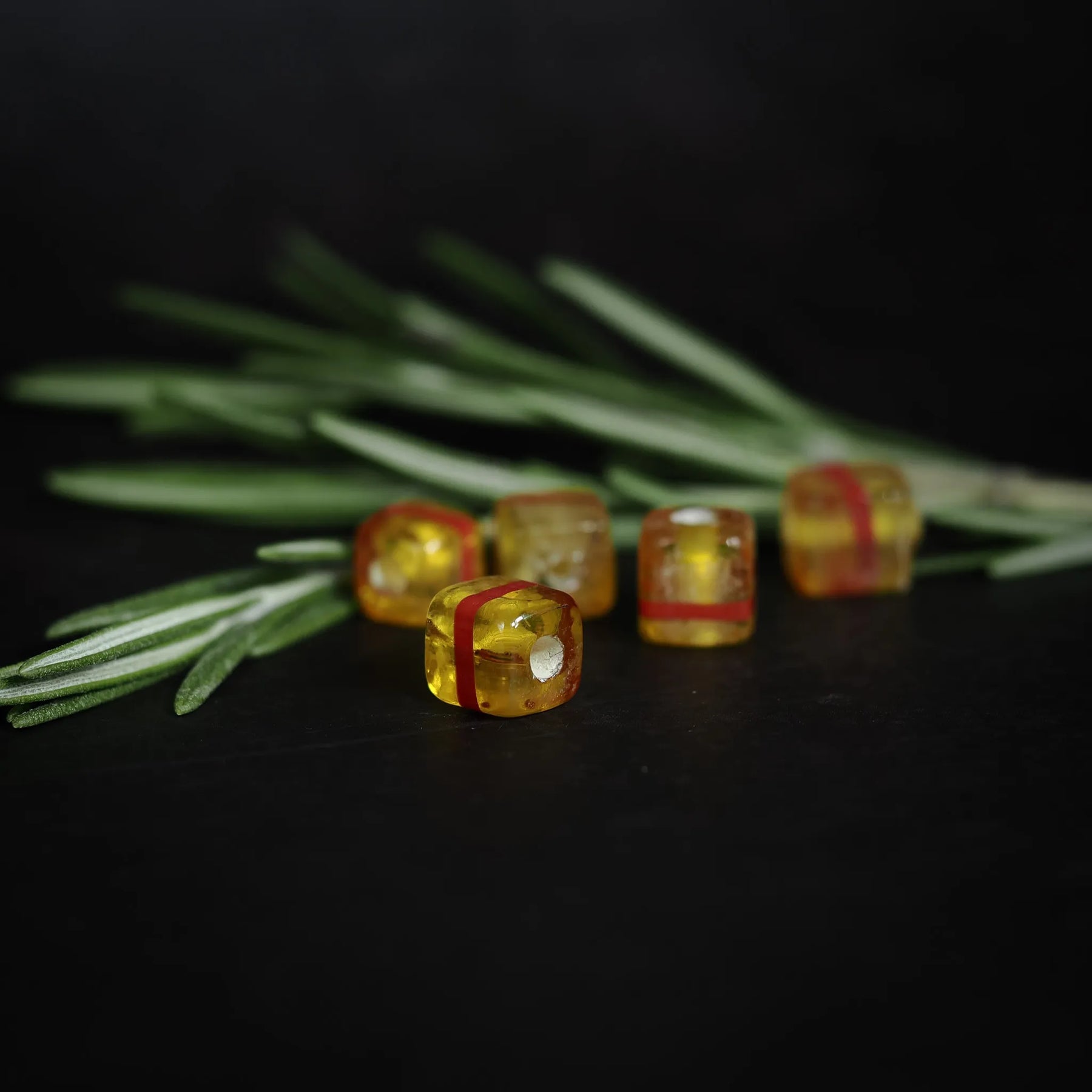 Fem gula kubiska glaspärlor med röda ränder på rad mot mörk bakgrund med barrnål, visar 9mm storlek och handgjorda detaljer