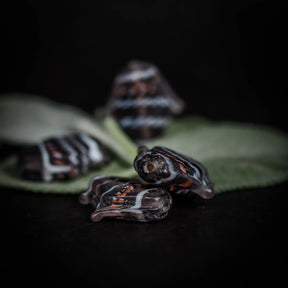 Handgjord glaspärla i form av fisk, grålila färg med rödvita mönster. Cirka 23x20x5mm med 1mm hål för tråd. Fotograferad på grön löv.