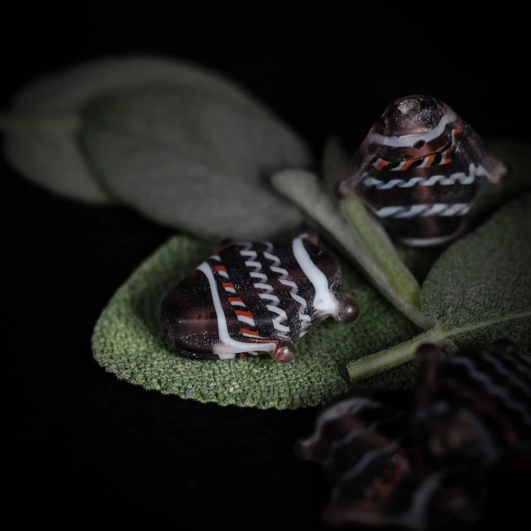 Handgjord glaspärla i form av fisk, grålila med rödvita strimmor. 23x20x5mm med 1mm hål. Närbild på grön löv med suddig bakgrund.