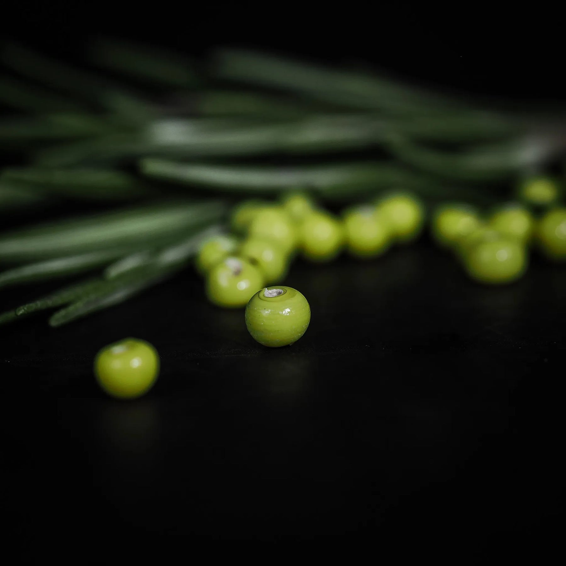 Närbild av handgjord limegrön glaspärla, 4mm i diameter, med hål för fiskelina. Glans glasyta, fotograferad på barrgren.