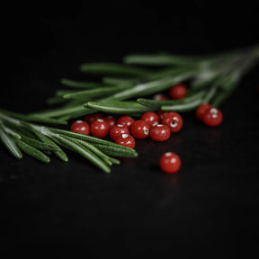 Handgjorda små röda glaspärlor, cirka 5mm diameter, med hål för fiskelina. Liggande på gröna barrgrenar mot mörk bakgrund.