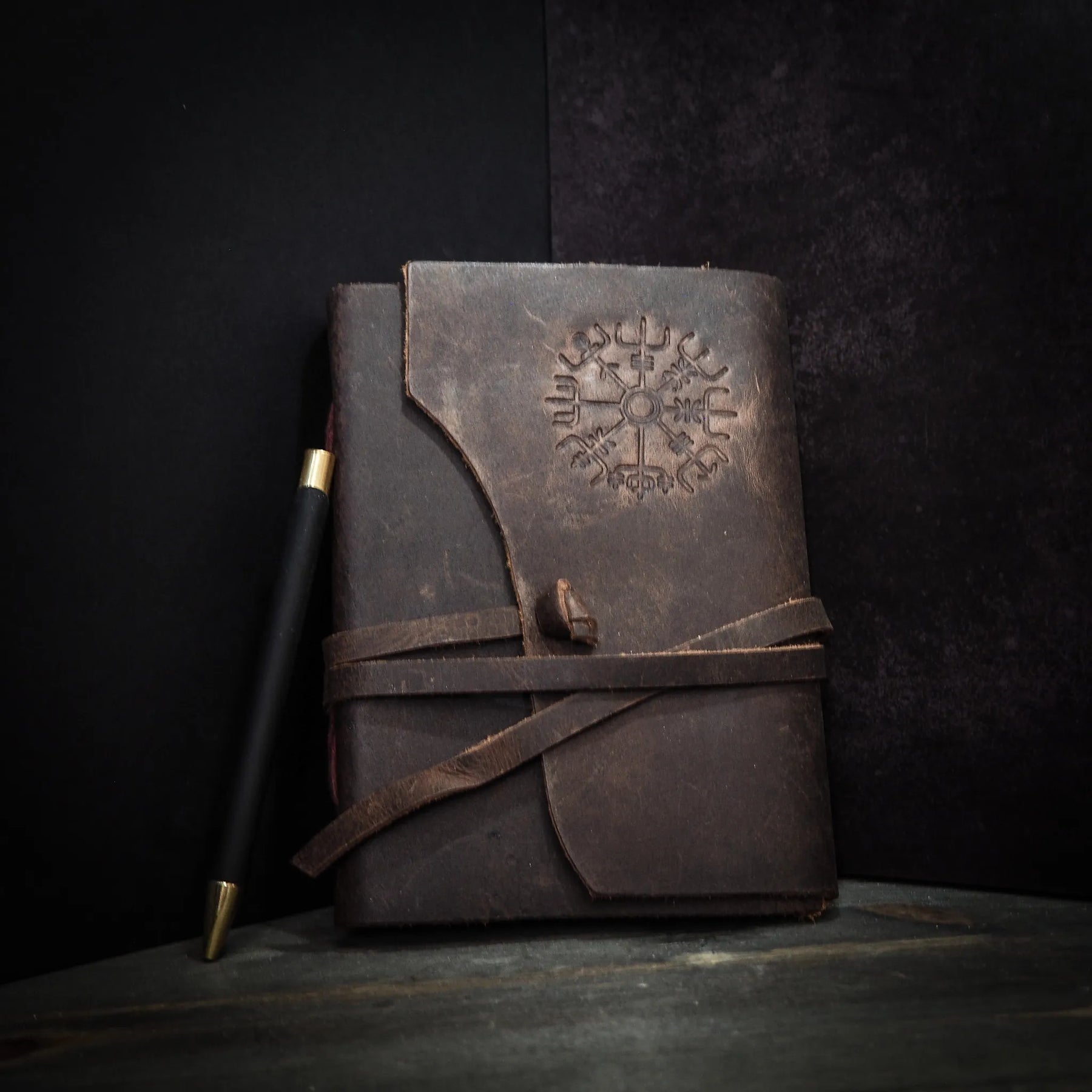 Leather journal with Vegvisir embossed symbol, shown at angle with writing pen for scale reference
