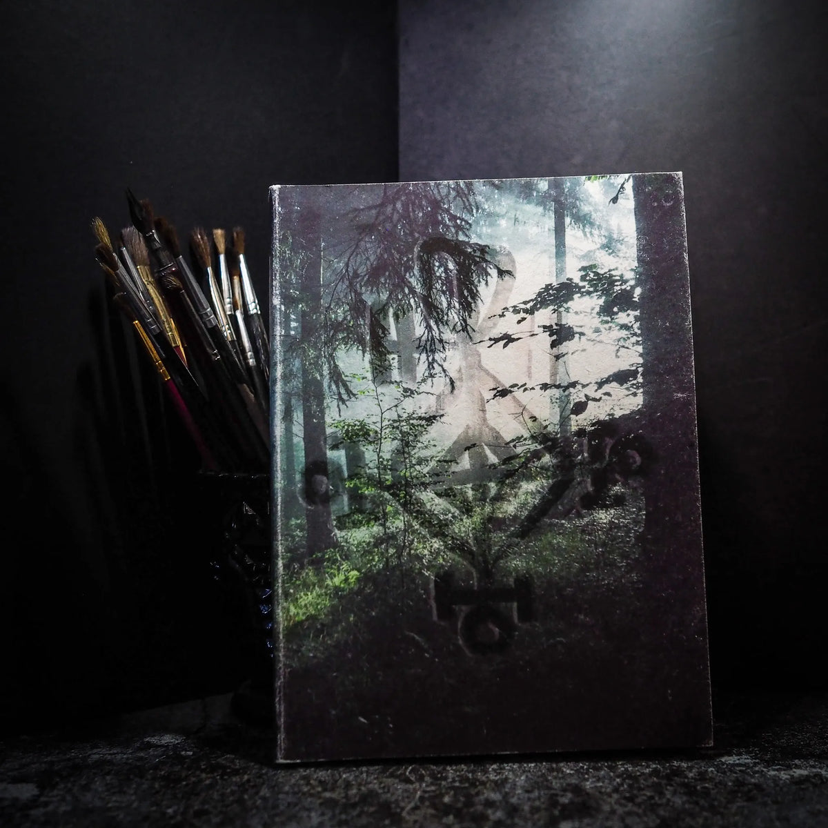 A5-målarbok med kärleksruna-motiv och skogsbakgrund, omgiven av konstnärspenslar och målarmaterial på mörk bakgrund.