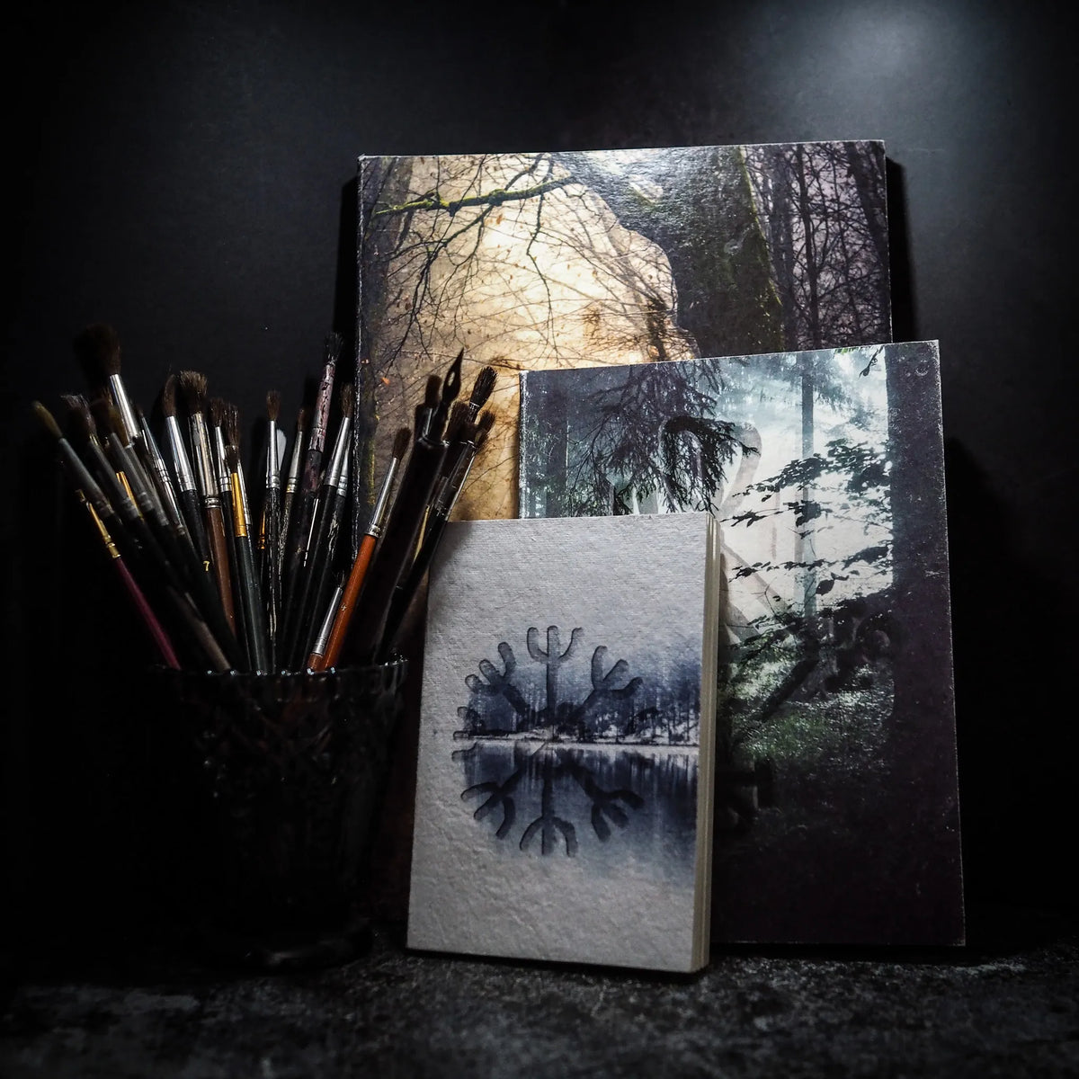 Två A5-målarböcker i konstnarlig stilleben tillsammans med penslar och målarverktyg, visar skogs- och naturmotiv.
