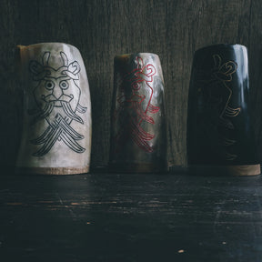 Three horn drinking mugs side by side with Odin mask engravings in different color variations - light cream horn with black, natural horn with red, and dark horn with cream engravings