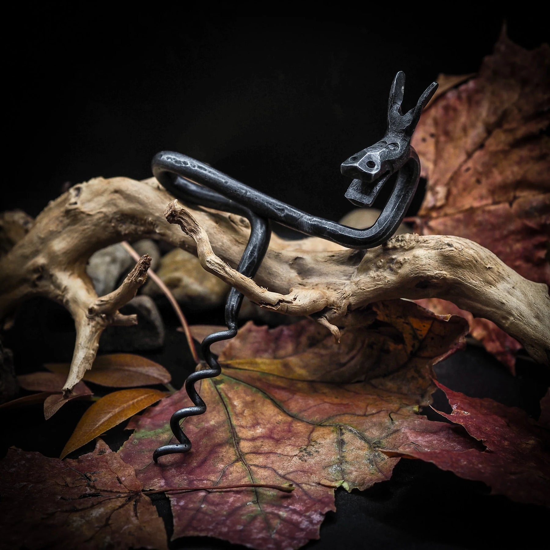 Älgskruv styld med höstlöv och grenar på naturlig bakgrund. Svart metall kontrasterar mot gyllene och bruna höstfärger. Älghuvudet och hornen är tydliga. Skapar höstlig skogshumör och jägar-stugans atmosfär. Betonar objektets dekorativa samlarkaraktär.