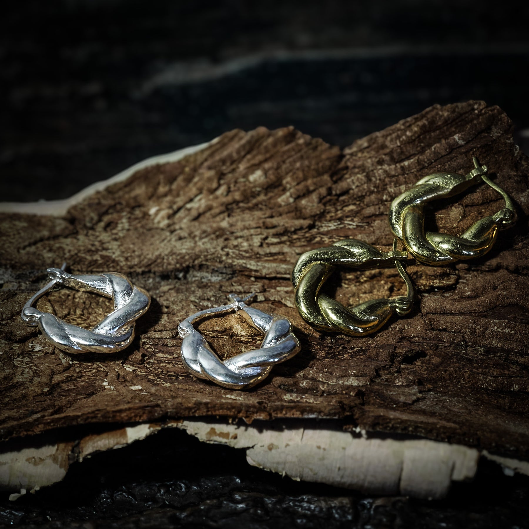 Flera par örhängen i flätad mässing på naturlig trägren. Visar både silver- och guldöverlagring, cirka 2 cm stora