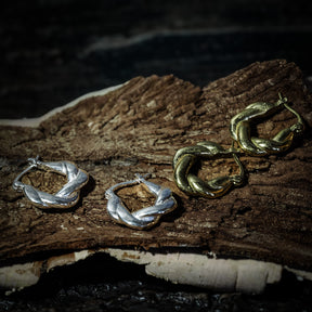 Flera par örhängen i flätad mässing på naturlig trägren. Visar både silver- och guldöverlagring, cirka 2 cm stora