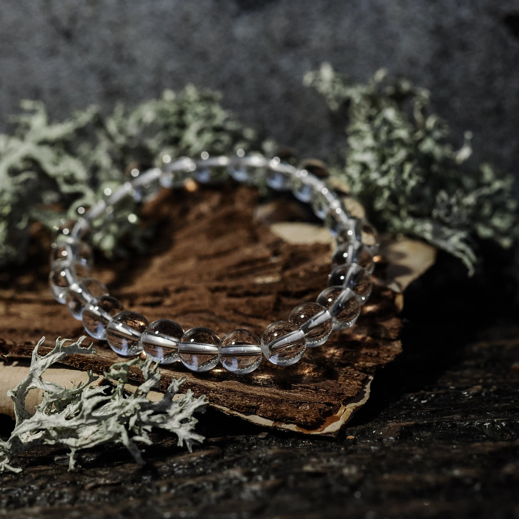 Armband stenpärlor - Tigeröga variant med bergkristall. Elastiskt armband med klara polerade kristallpärlor som speglar ljus, på rustik träbas med mossbeväxt stenbackgrund.