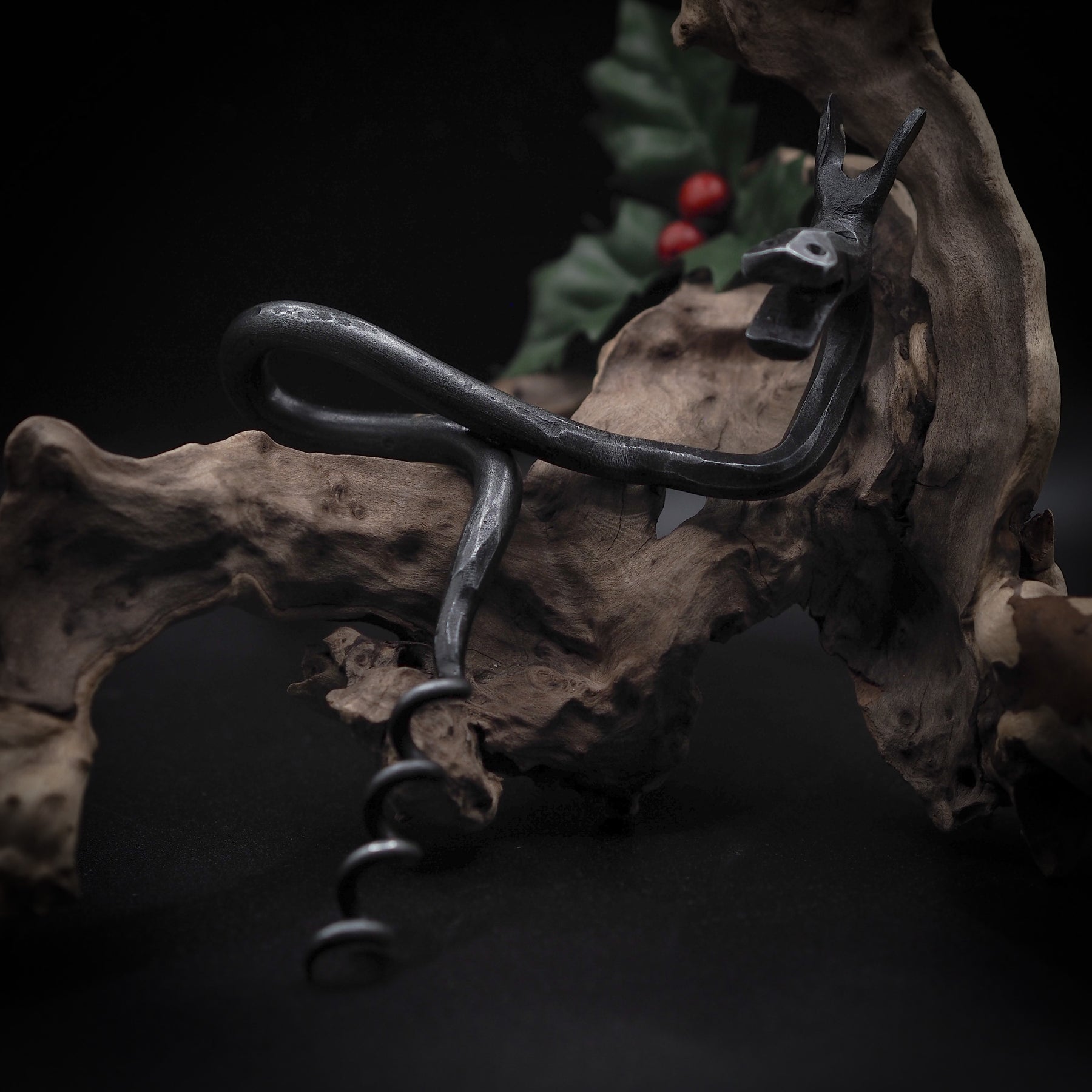 Älgskruv på väderbitven trägren i skogsliknande miljö. Älghuvudet visar framträdande horn. Suddiga röda bär och grön vegetation i bakgrunden. Mörkmetallen kontrasterar mot naturligt trä. Atmosfärisk belysning betonar skulpturens tredimensionella form.