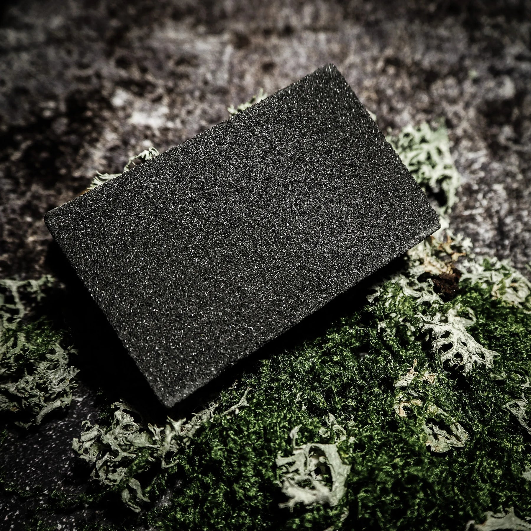 Dark grey rectangular abrasive cleaning block for rust removal, shown on moss and stone background
