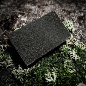 Dark grey rectangular abrasive cleaning block for rust removal, shown on moss and stone background