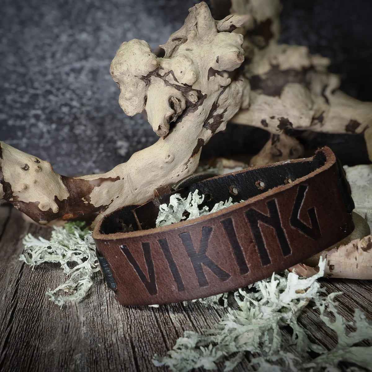 Brunt läderarmband med graverade vikingruna som läser 'VIKING', presenterad med naturliga rekvisita av trä och lav