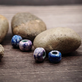 Fyra blålila glasspärlor med olika mönster (vita prickar, blandade färger), fotograferade på trä med naturliga stenar