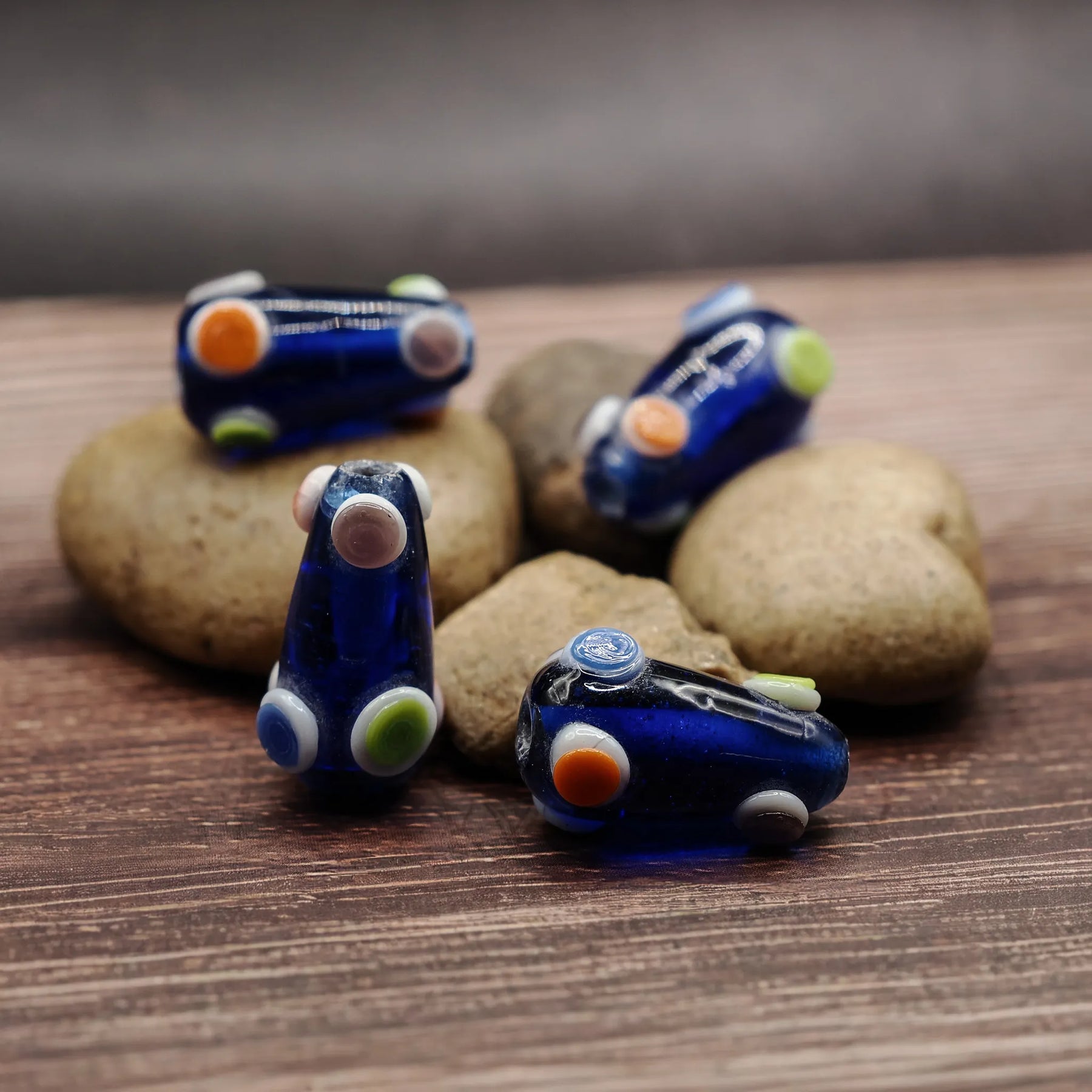 Fem mörkblå formade glasspärlor med färgade prickar (orange, gul, grön, vit), fotograferade på trä med stenar