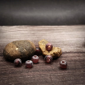 Mauve-colored glass beads, small spherical beads approximately 5 millimeters in diameter. Handmade beads in muted purple-grey tone with matte finish. Several beads shown on natural wooden base with visible holes for threading.
