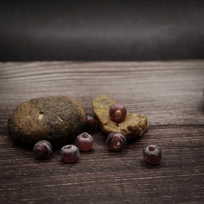 Mauve-colored glass beads, small spherical beads around 5 millimeters. Handmade beads with muted purple color and matte finish. Several beads arranged on natural wooden surface showing artisanal characteristics.