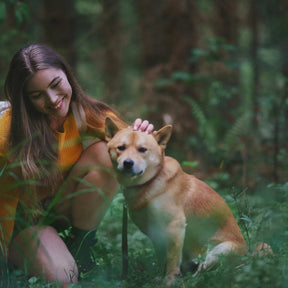 Livsstilsbild av kvinna i gul utomhuskläder med brun hund i skogsmiljö. Titeln 'Kalla på hunden' visar visselpipans praktiska användning som hundsignal under vandring och outdoor-aktiviteter.